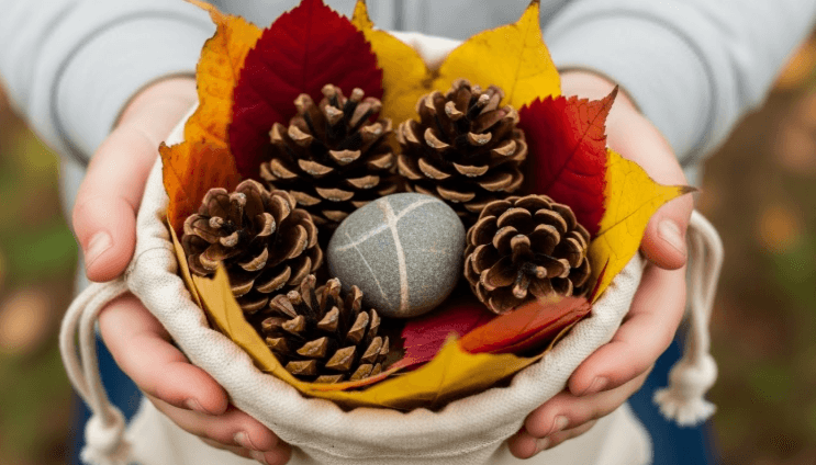 Close-up of a child's hands holding a small canvas bag filled with pinecones, colorful leaves, and a smooth river stone]