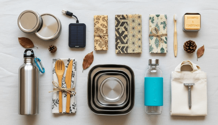 Flat lay of zero waste camping essentials including stainless steel containers, bamboo utensils, beeswax wraps, and reusable water bottles arranged on a neutral background